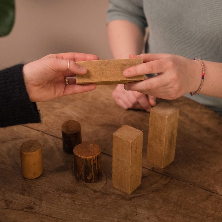 Hände tauschen einen Holzblock über einem Tisch mit geometrischen Holzformen.