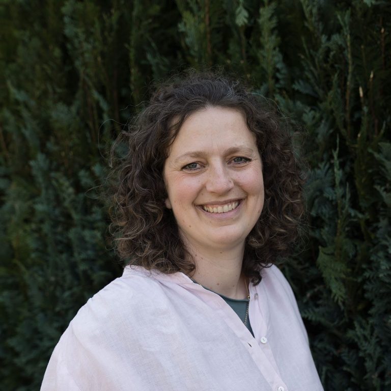 Freundliche Frau mit lockigem Haar vor grünen Pflanzen.