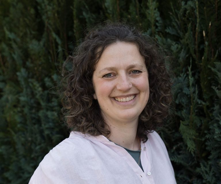 Junge Frau mit lockigem Haar, lächelt vor einem Hintergrund aus grünen Pflanzen.