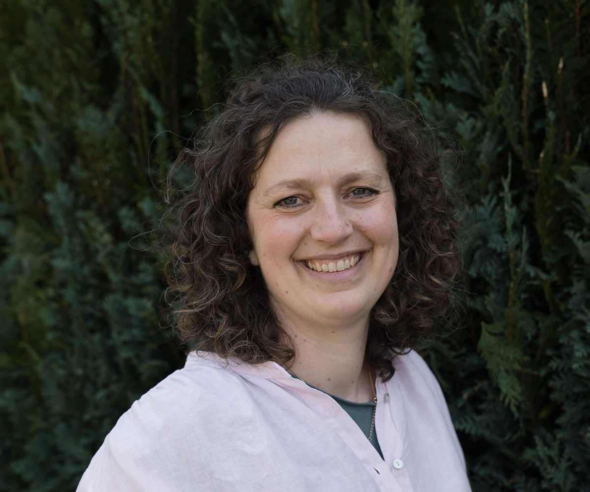 Junge Frau mit lockigem Haar, lächelt vor einem Hintergrund aus grünen Pflanzen.