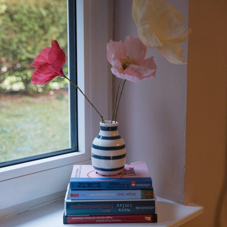 Bunte Blumen in einer gestreiften Vase auf einem Stapel Bücher am Fenster. Titel von Jesper Juul und Nora Imlau.