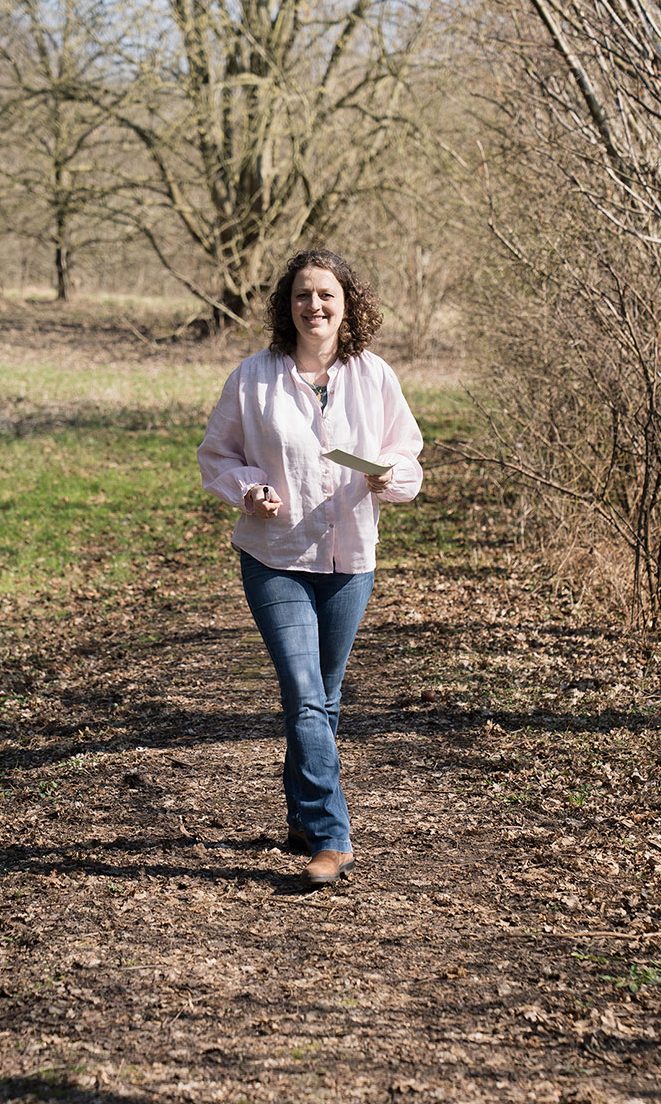 Eine Frau mit Karten in der Hand läuft einen Weg entlang, umgeben von Bäumen.